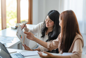 Two women sitting together looking at a clipboard, on which there's a document with project management statistic infographics on it, One is mentoring the other as a project management apprentice.